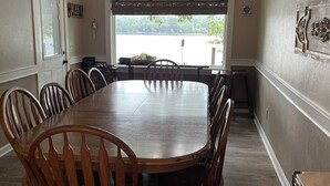 Comedor con gran mesa y sillas de madera, ventilador de techo y vista a una masa de agua a través de amplias ventanas.