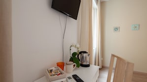 A room with a flat-screen TV mounted on the wall, a coffee maker, a remote control, a mug, and a small plant on a white table. - Suites Colombo 32 (La Spezia)