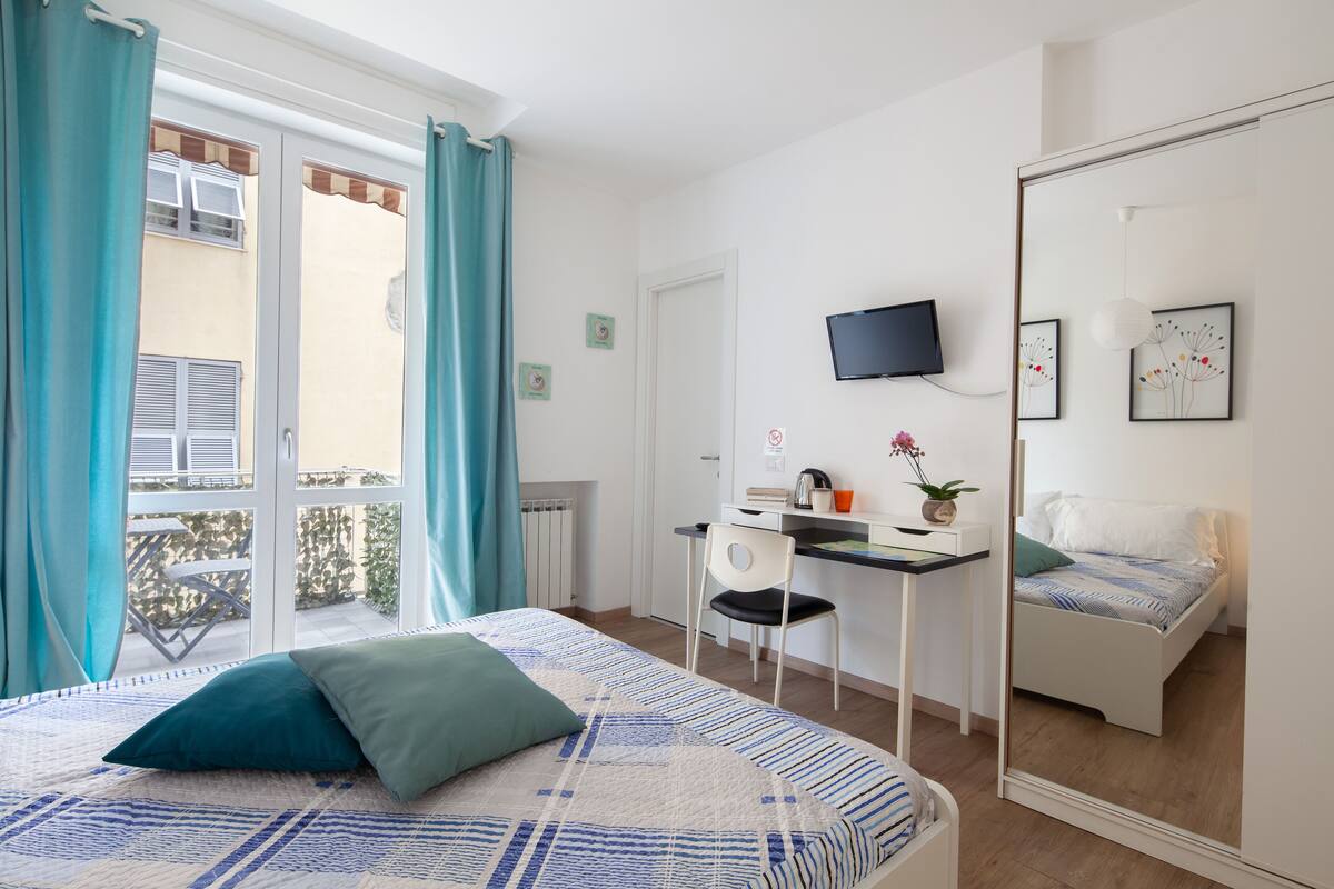 Quarto com cama, escrivaninha, cadeira, televisão e varanda com cortinas.