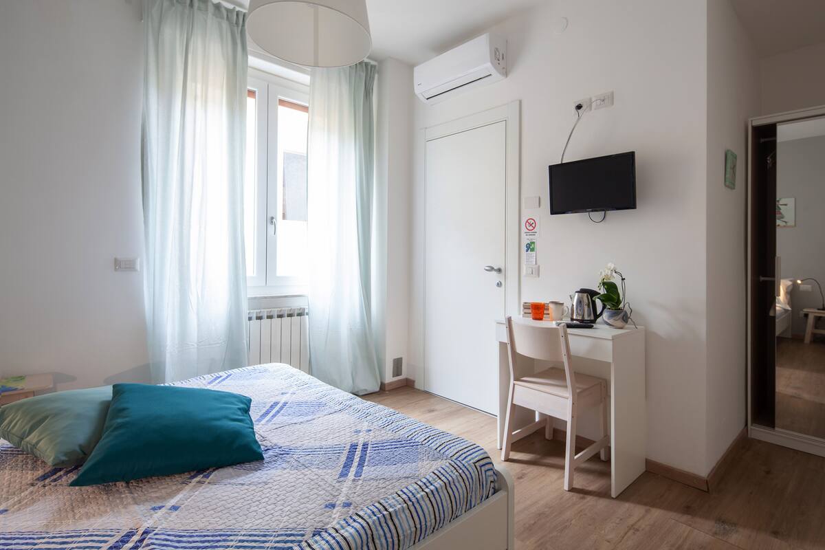 Quarto com cama, escrivaninha, cadeira, televisão e janela com cortinas.