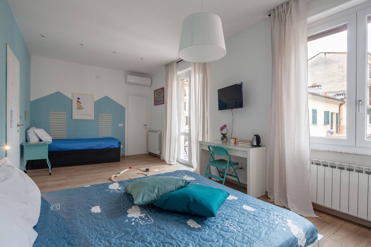 Quarto com cama, escrivaninha e cadeira. Tem uma janela com cortinas, radiador e televisão na parede.