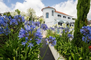 A white building with a red roof, surrounded by a garden with blue and white flowers. - Blue Sea Tower, Luxury Villa, Private Heated Pool and Gardens. Superb Ocean view (Fajã da Ovelha)