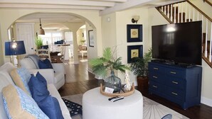 A living room with a white sofa, a round coffee table, a TV on a blue stand, and a staircase in the background. - Sunny Claire- Beautiful In Town Cottage w/ Hot Tub (Chincoteague)
