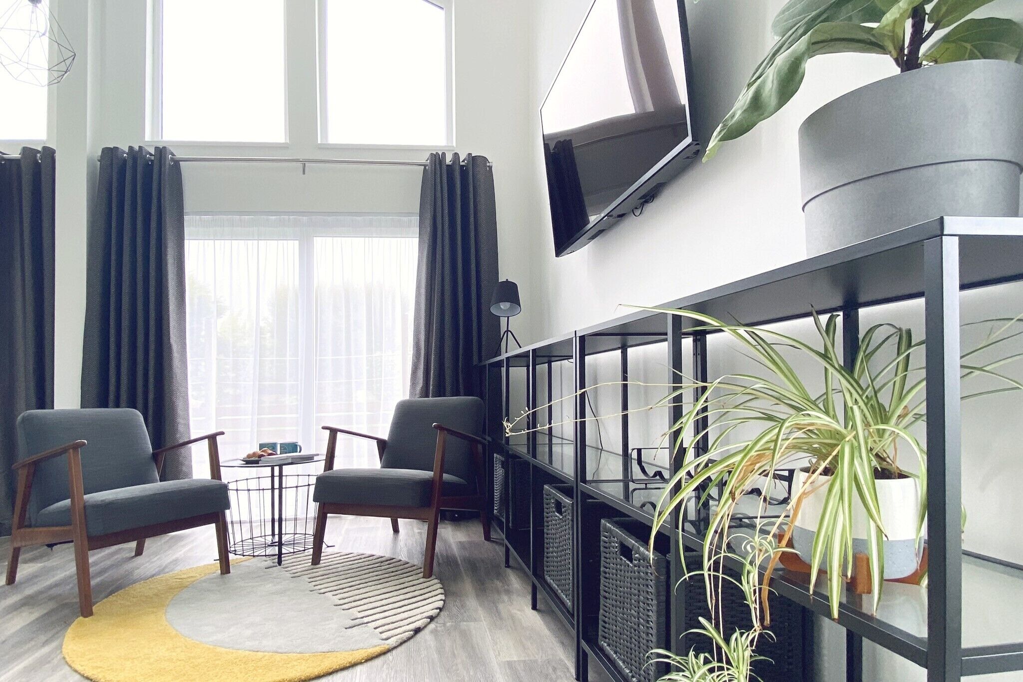 A modern living room with two grey armchairs, a small round table, a yellow rug, and a black console table with plants.