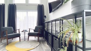 Ein modernes Wohnzimmer mit zwei grauen Sesseln, einem kleinen runden Tisch, einem gelben Teppich und einer schwarzen Ablagetisch mit Pflanzen.