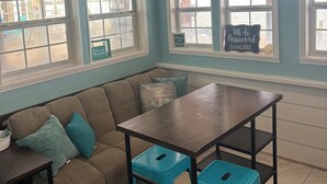Une pièce avec un canapé marron, une table en bois et deux tabourets bleus. Il y a des fenêtres avec des cadres blancs et une enseigne sur le mur qui indique : « SALTY AIR DR ».