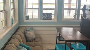 Une pièce avec un mur bleu, une enseigne indiquant « SALTY AIR DR », une fenêtre donnant sur l’eau, un canapé marron avec des coussins turquoise, une table en bois et deux tabourets bleus.
