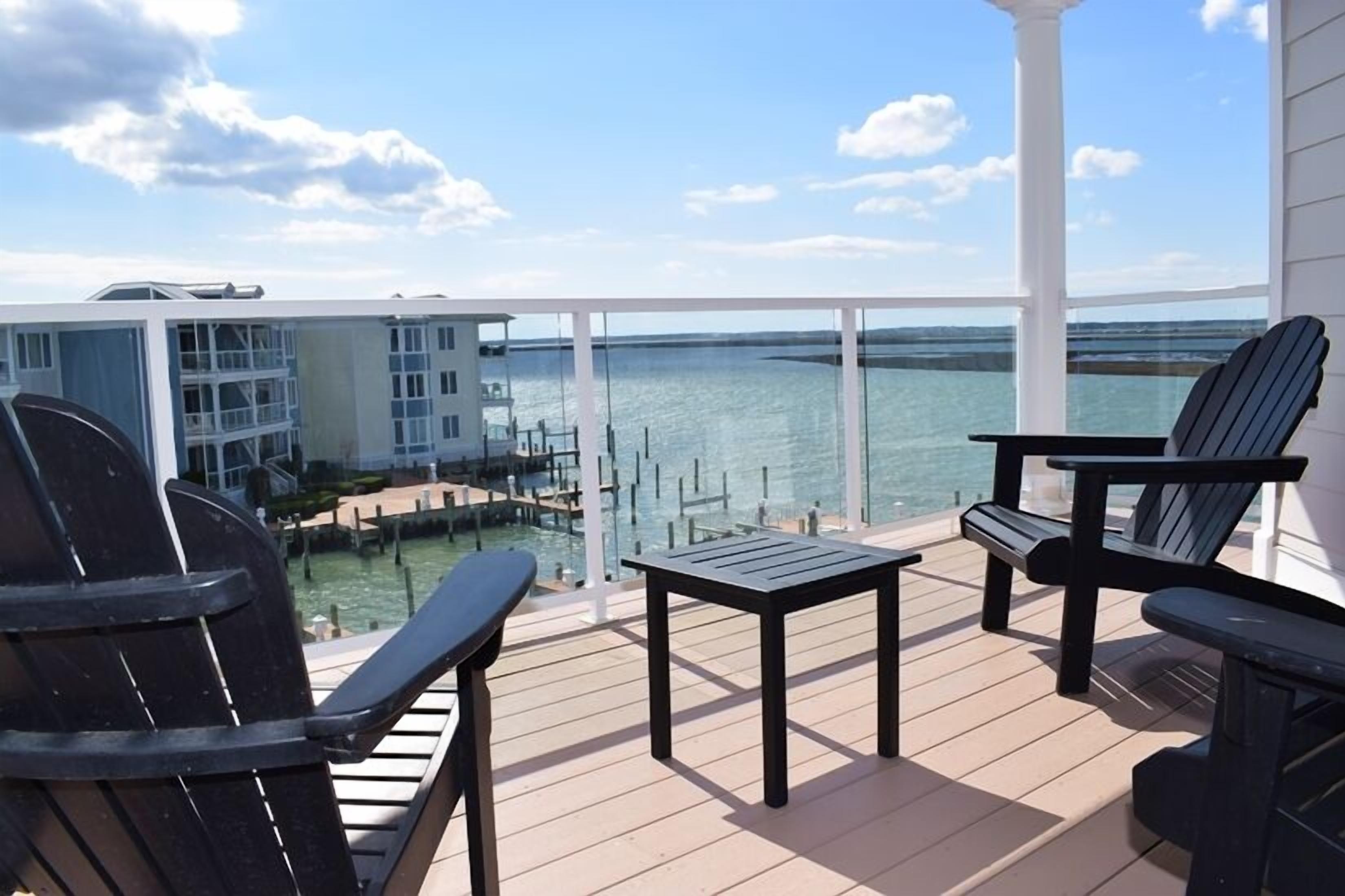 Une terrasse avec deux fauteuils Adirondack et une petite table, donnant sur une marina où sont amarrés des bateaux, avec un bâtiment visible au loin.
