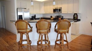 Una cocina con gabinetes blancos, un refrigerador de acero inoxidable, un microondas e isla de madera con tres taburetes de mimbre.