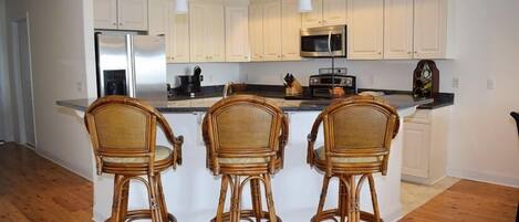 A kitchen with white cabinets, a stainless steel refrigerator, a microwave, and a wooden island with three wicker bar stools.