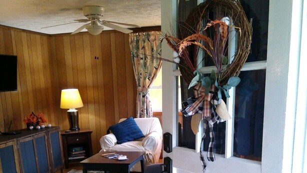 Un salon avec des murs en bois, un ventilateur de plafond, un canapé avec un plaid bleu, une table basse, une lampe, une décoration en osier et une porte avec une fenêtre.