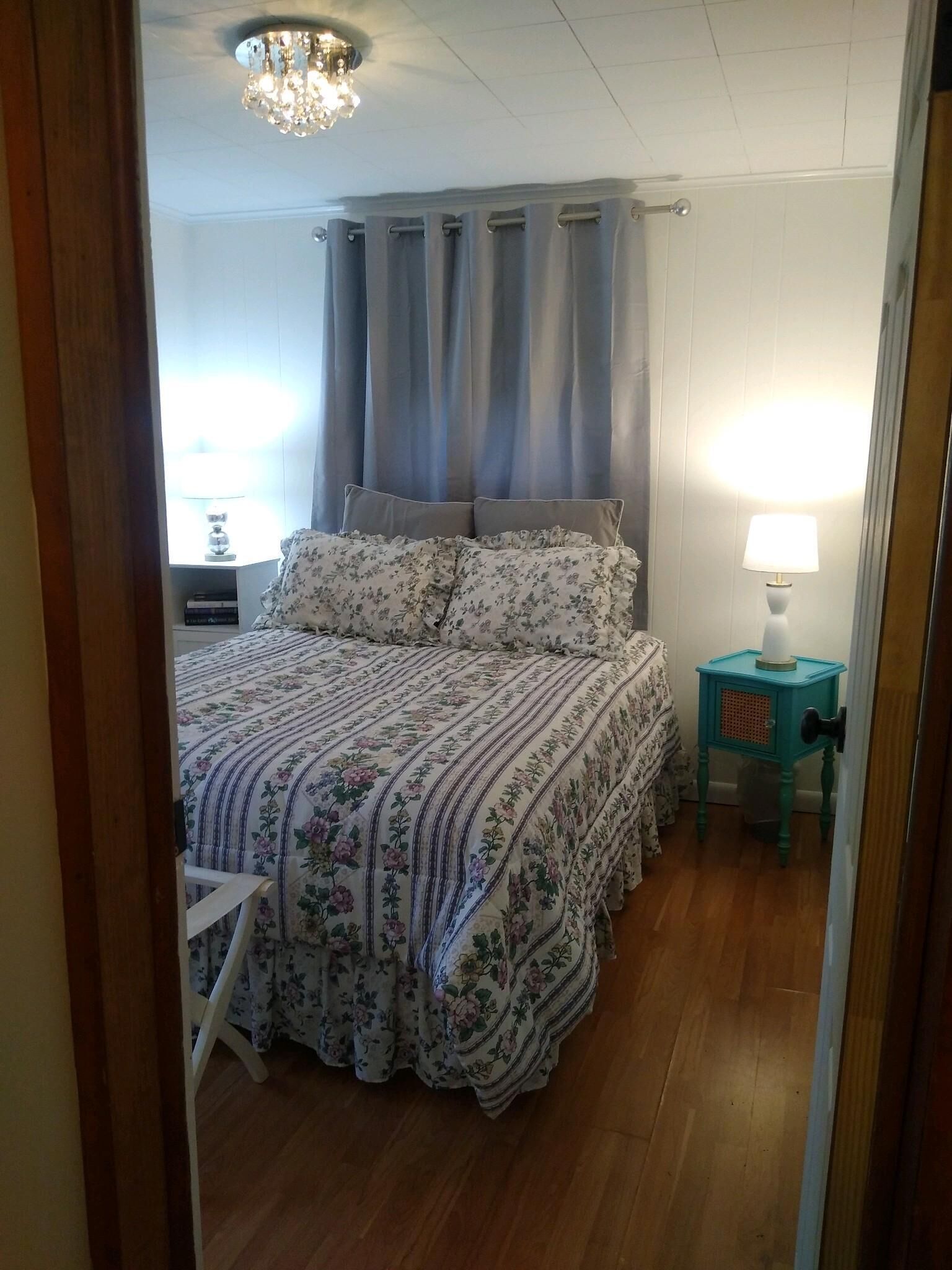 Un dormitorio con una cama, cortinas, una mesita de noche, una lámpara y suelo de madera.