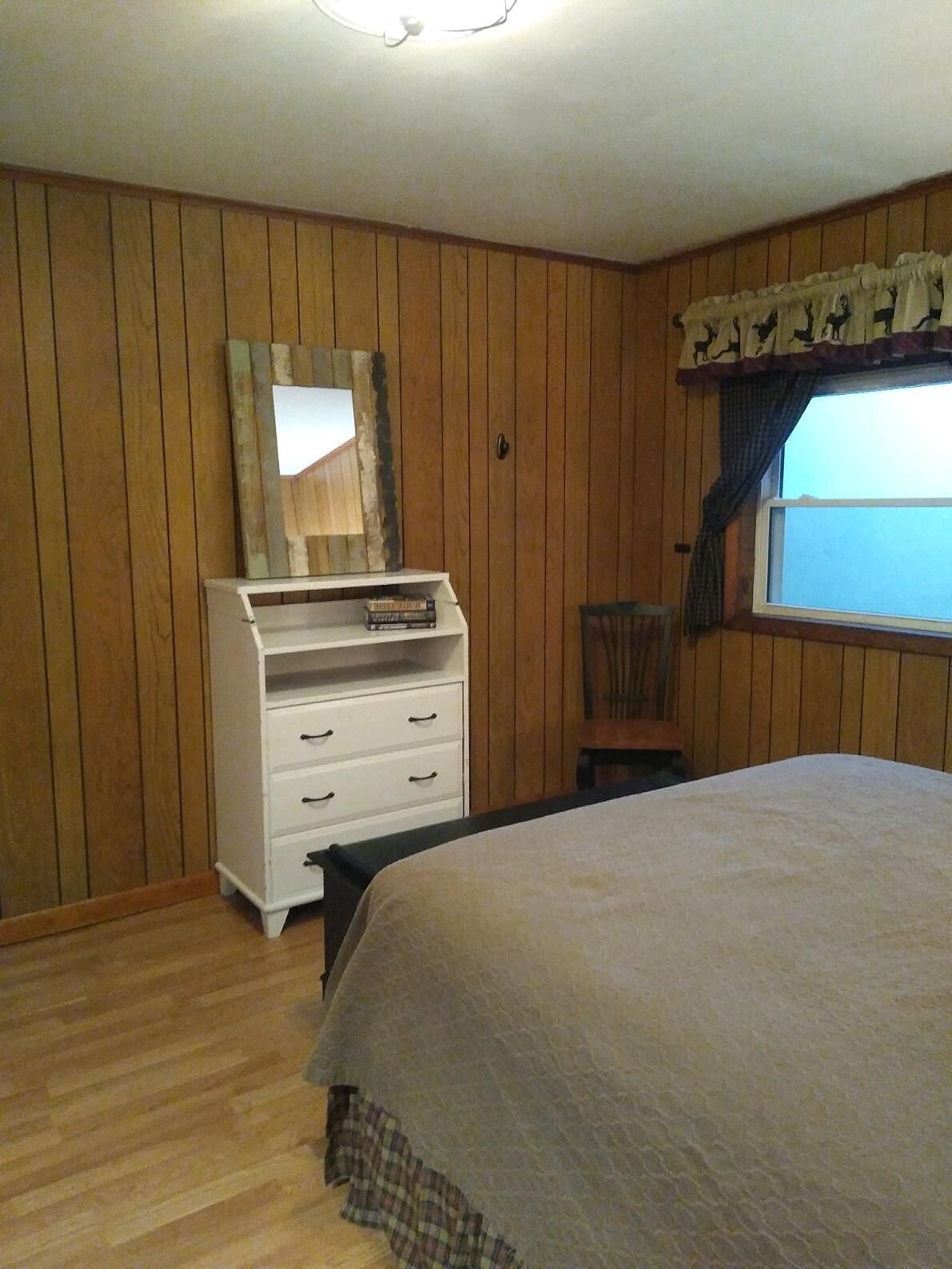 Une chambre avec des lambris en bois, un lit, une commode à tiroirs, une chaise et une fenêtre avec des rideaux.