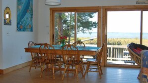 Un coin repas avec une table et des chaises en bois, un vase rempli de fleurs et une vue sur la mer grâce à de grandes fenêtres.