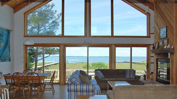 Une pièce spacieuse avec un plafond haut, de grandes fenêtres offrant une vue sur la mer, un coin repas avec des meubles en bois et un coin salon confortable agrémenté de coussins à rayures.