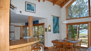 Un vaste espace dédié à la salle à manger, avec une table et des chaises en bois, un bar avec des tabourets, et de grandes fenêtres offrant une vue sur les arbres.