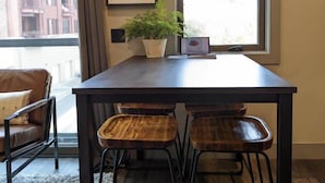 Un coin repas comprenant une table, quatre chaises en bois, une plante en pot, un tableau encadré et une fenêtre avec des stores.