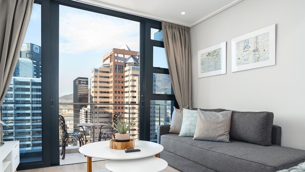 Un salón moderno con un sofá gris, una mesa de centro blanca y vistas de edificios de la ciudad a través de amplias ventanas.