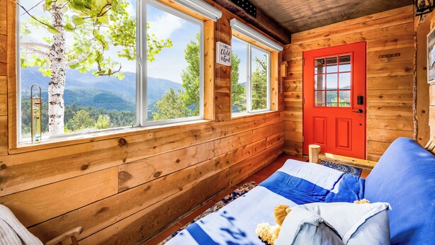 A cozy cabin room with wooden walls, a red door, and large windows offering a mountain view.