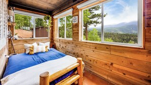 A wooden cabin room with a bed, large windows, and a view of a forested landscape.