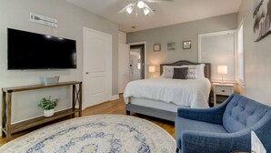 A bedroom with a bed, a TV, a ceiling fan, a bedside table, a chair, and a window with blinds.