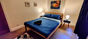 A neatly made bed with a blue bedspread, two bedside lamps, and a painting of a sunset on the wall.