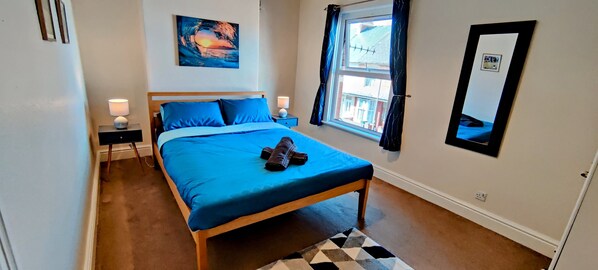 A bedroom with a bed, two bedside tables with lamps, a window with curtains, a wall-mounted mirror, and a painting.