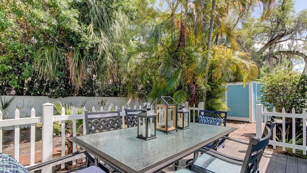 Un patio exterior con mesa y sillas, rodeado por una valla de piquetes blancos y abundante vegetación.