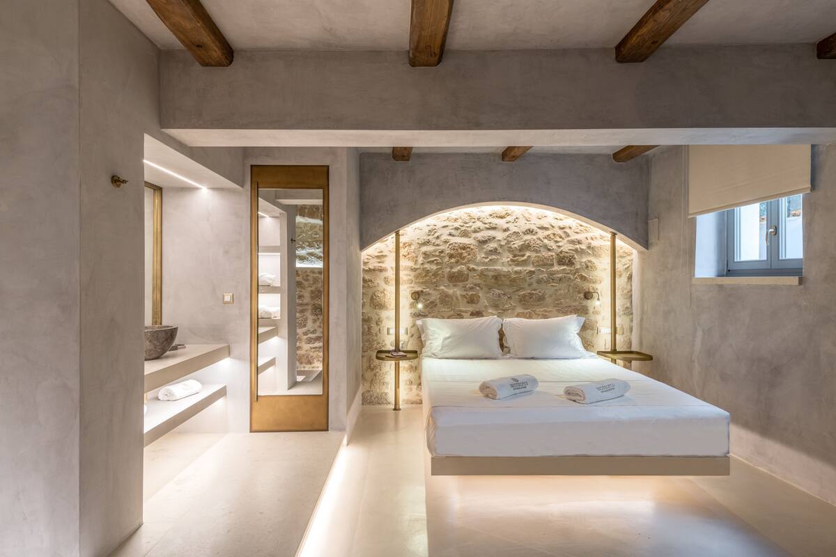 Une chambre avec une arche en pierre, un lit recouvert de draps blancs, une table de chevet et une fenêtre donnant sur un paysage agréable.