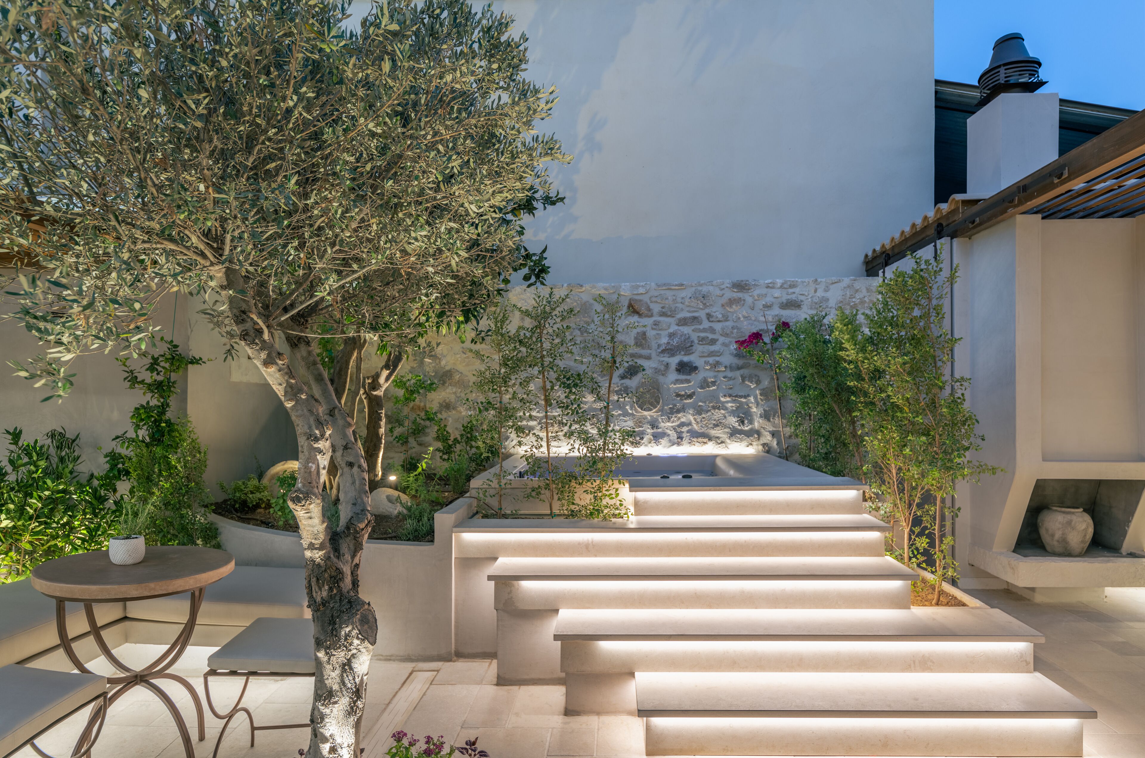Un espacio exterior moderno con un muro de piedra, un árbol y un conjunto de escalones que conducen a una zona de estar.