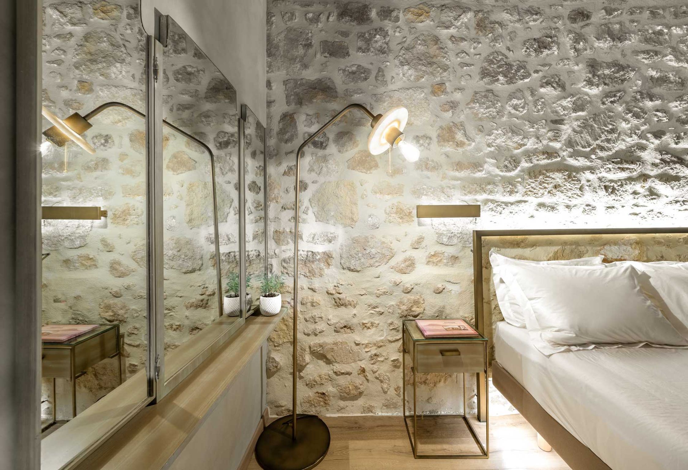 A bedroom with a stone wall, a bed with white linens, a bedside table with a lamp, and a mirror.