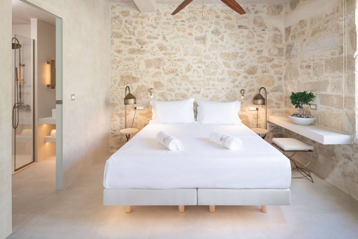 Une chambre avec un mur en pierre, un lit recouvert de draps blancs, deux lampes de chevet et une plante en pot.