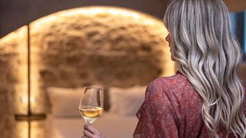 A person holding a glass of white wine in a room with a stone wall and a bed in the background.