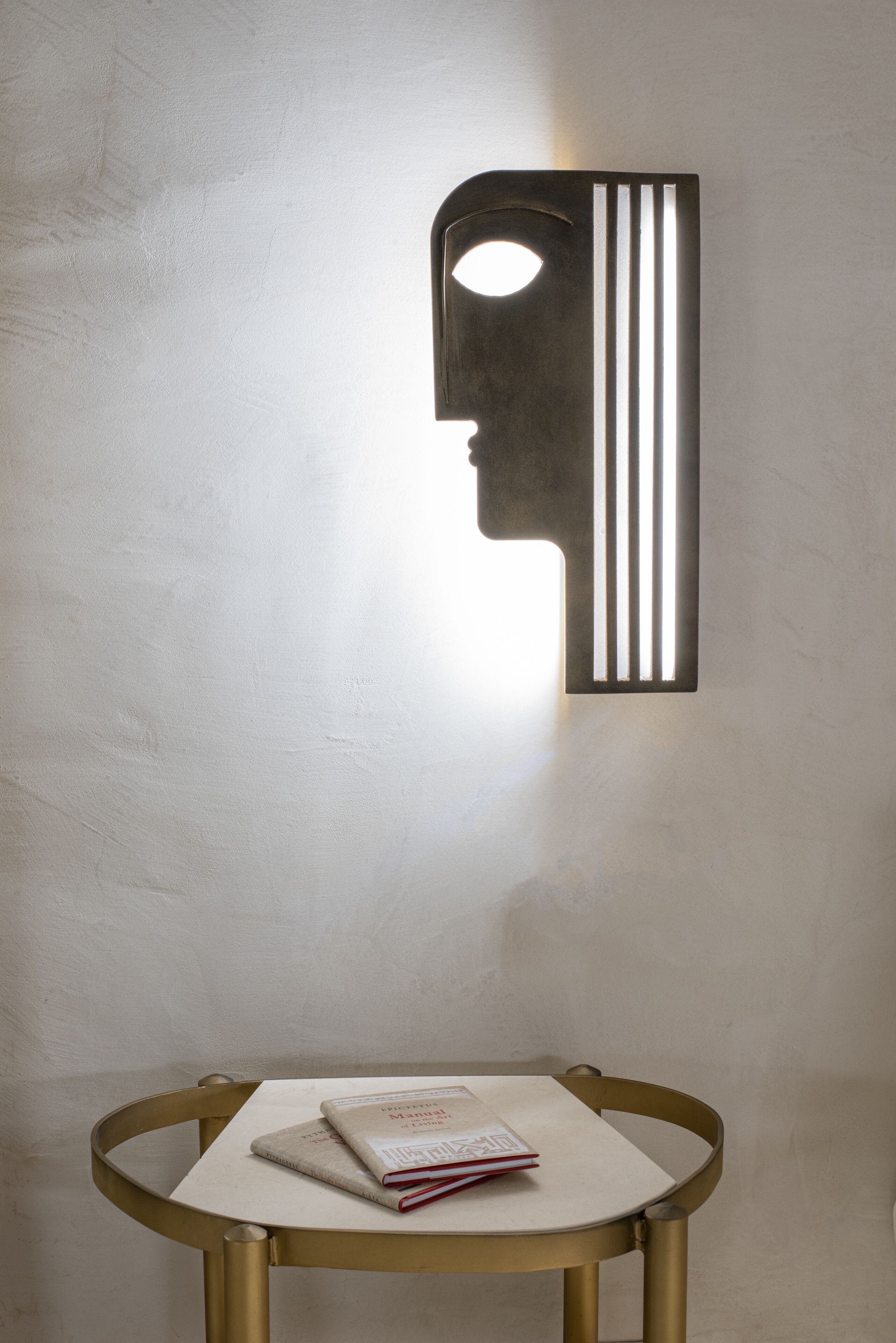 Una lámpara de pared con diseño geométrico, una mesita con un libro y una silla.