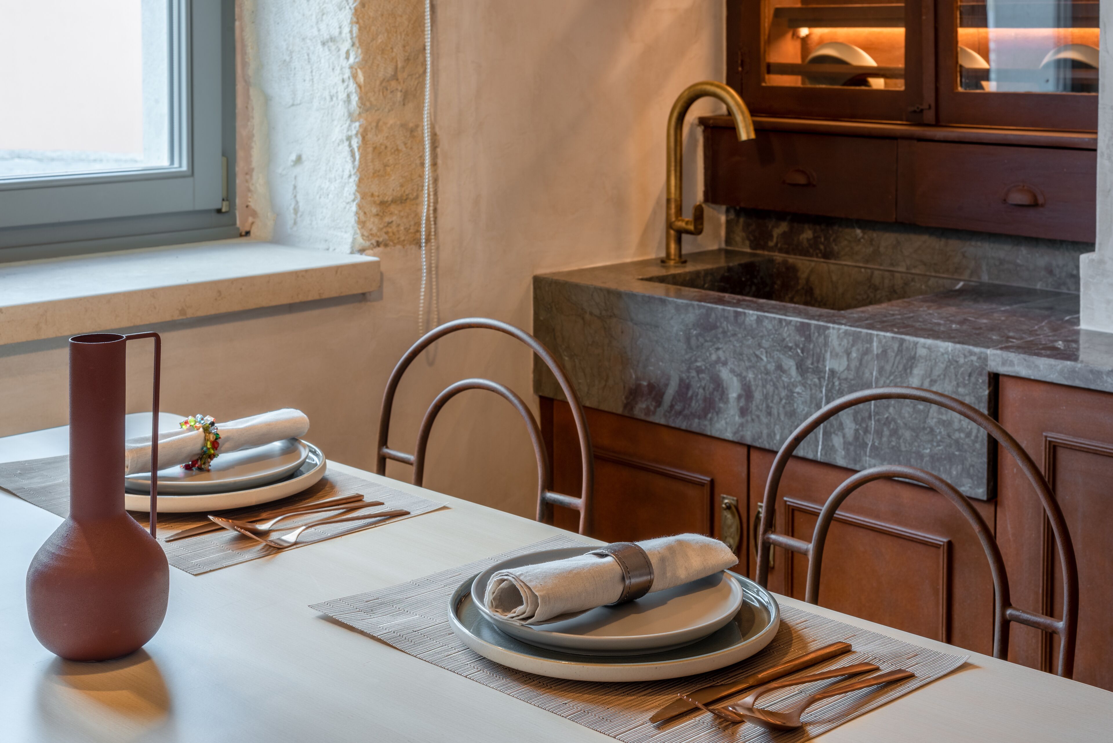 Un coin repas avec une table dressée pour deux personnes, un vase et une cuisine équipée d’un évier et de placards.