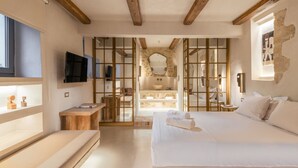 A modern bedroom with a large bed, wooden beams, a TV, a wooden bench, and a bathroom with a stone wall and bathtub. - Inotropes Boutique Suites - Adults Only (Archanes-Asterousia)