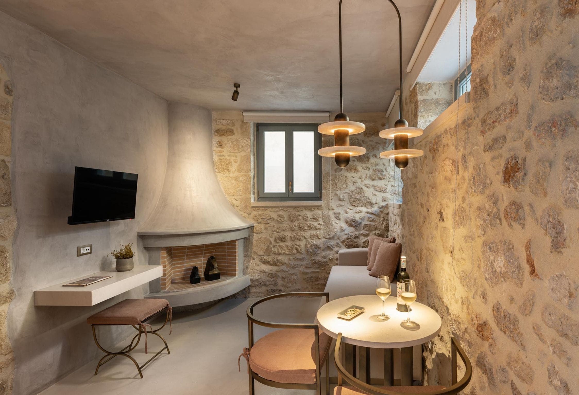 A cozy interior with a stone wall, a fireplace, a flat-screen TV, a round dining table with chairs, and pendant lighting.