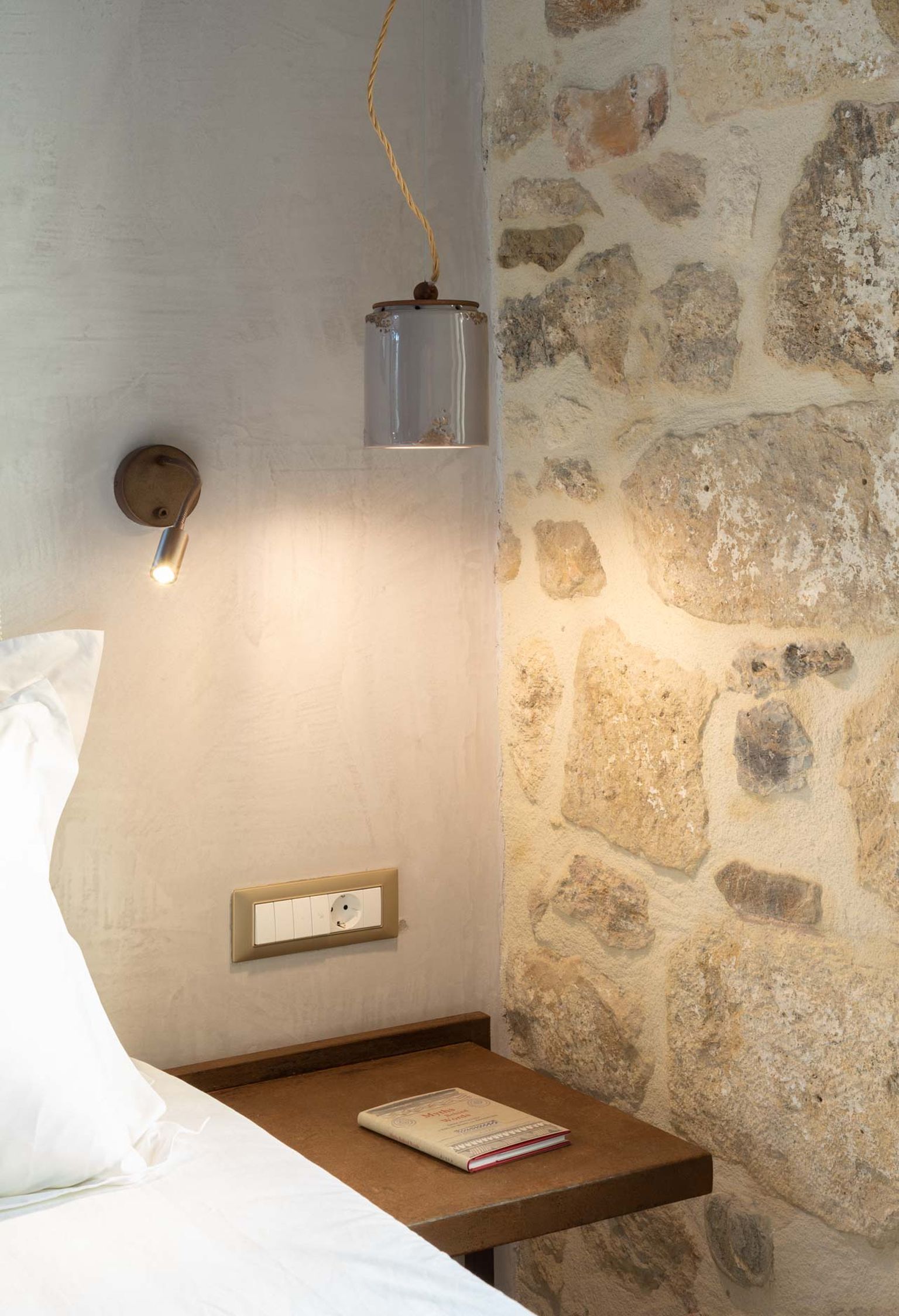 Camera da letto con parete in pietra, lampada da comodino, comodino e un libro.