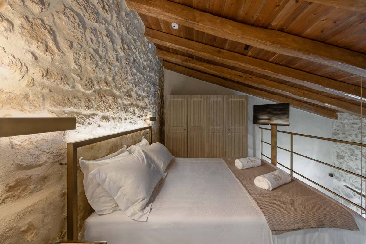 Une chambre avec un mur en pierre, des poutres en bois au plafond, un lit avec du linge de maison blanc, une armoire en bois et un téléviseur fixé au mur.