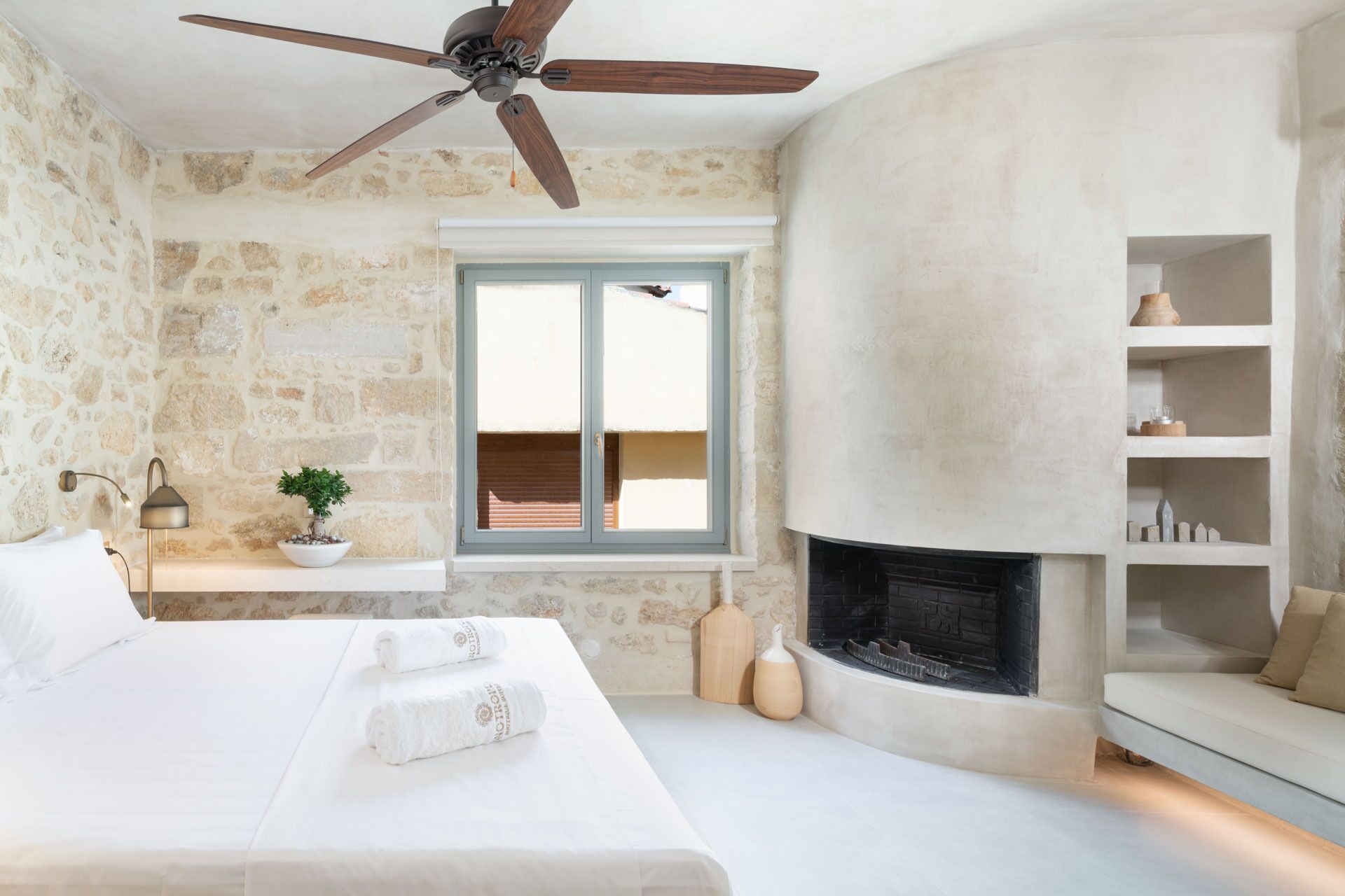 Um quarto com parede de pedra, cama com roupa de cama branca, ventilador de teto, janela com vista, lareira e prateleira com objetos decorativos.