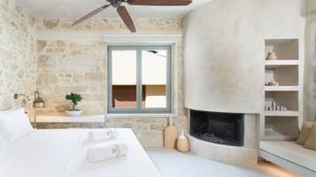 Um quarto com parede de pedra, cama com roupa de cama branca, ventilador de teto, janela com vista, lareira e prateleira com objetos decorativos.
