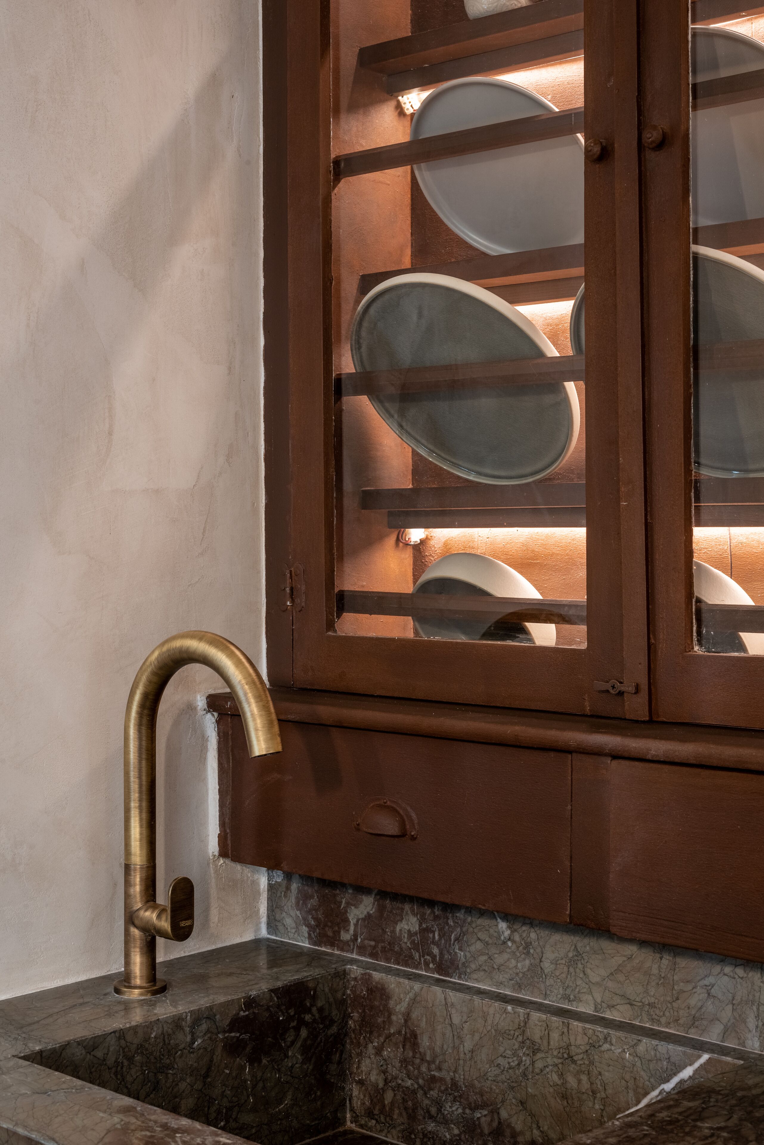 Un baño con un lavabo de mármol, un grifo de latón y un mueble de madera con puertas de cristal que contiene platos blancos.