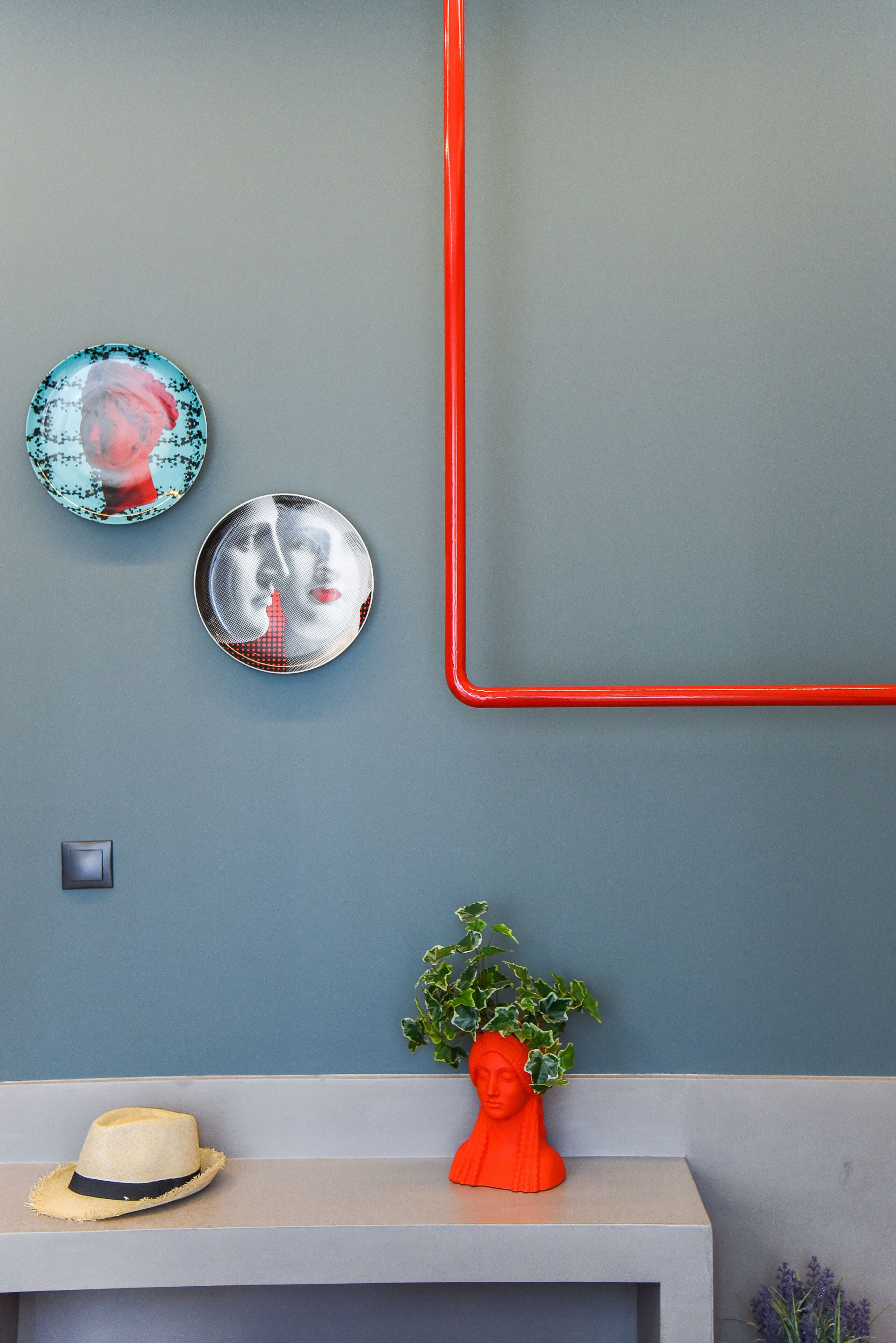 A wall with two decorative plates, a red pipe, a hat, and a plant on a shelf.