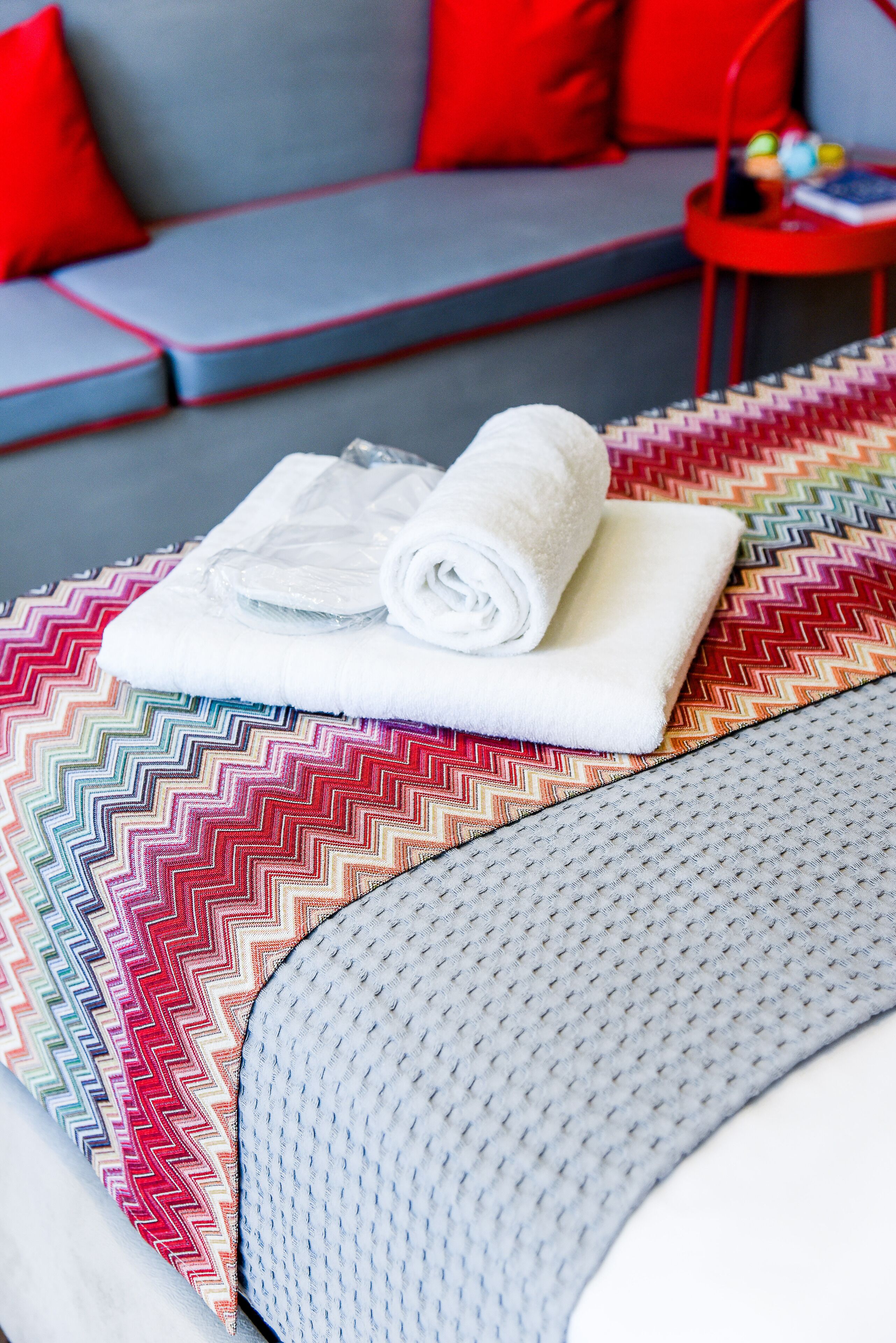 A neatly made bed with a rolled white towel and a transparent plastic bag, a colorful zigzag patterned rug, and a grey sofa with red cushions in the background.