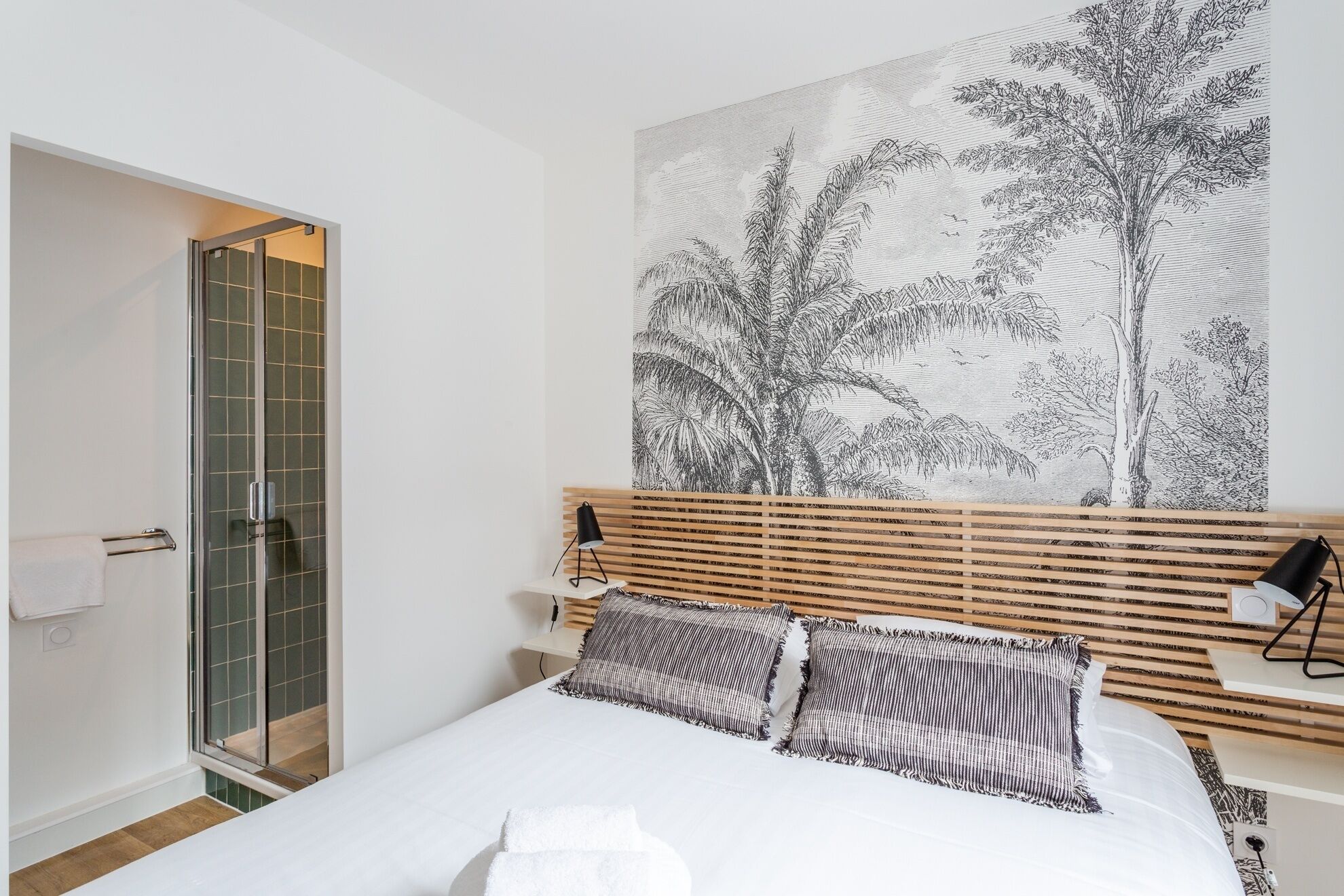 A bedroom with a bed, a wooden headboard, a large wall art of trees, a bedside table with a lamp, and a glass shower enclosure.