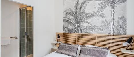 A bedroom with a bed, a wooden headboard, a large wall art of trees, a bedside table with a lamp, and a glass shower enclosure.