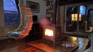 A cozy living room with a wood-burning stove, a wicker chair, and a bookshelf. - Waterfront House and Artist's Studio in the Woods (Kent)