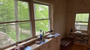 A cabin with a wooden interior, large windows overlooking a forest, a small desk with various items, and a bookshelf filled with books. - Waterfront House and Artist's Studio in the Woods (Kent)