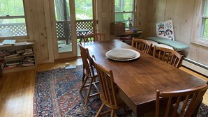 A wooden dining room with a large table, chairs, and a rug. There are bookshelves and a window with a view of trees. - Waterfront House and Artist's Studio in the Woods (Kent)