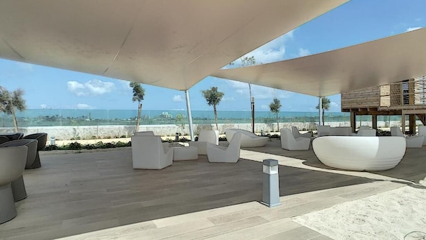 A modern outdoor seating area with white chairs and a wooden table, overlooking a beach. - Dreamy Sunset - Appartement (Playa Honda)